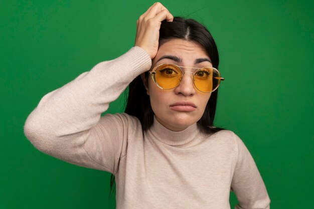 Confus jolie brune fille caucasienne dans des lunettes de soleil met la main sur la tête et regarde la caméra sur le vert