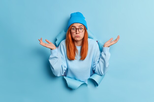 Confus et perplexe jeune femme interrogée aux cheveux rouges hausse les épaules et écarte les paumes sur le côté se tient sans aucune idée dans le trou déchiré du trou de papier bleu