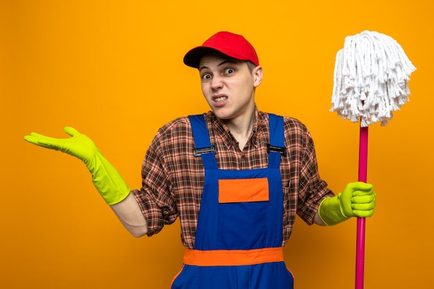 Confus de propagation des mains jeune homme de nettoyage portant un uniforme et une casquette avec des gants tenant une vadrouille