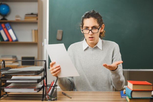 Photo gratuite confus répandre les mains jeune enseignant portant des lunettes tenant du papier assis au bureau avec des outils scolaires dans la salle de classe