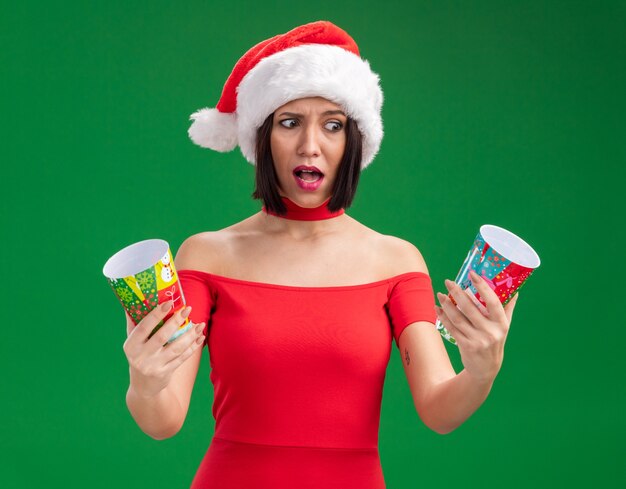 Confus young girl wearing santa hat holding gobelets de Noël en plastique à l'un isolé sur mur vert