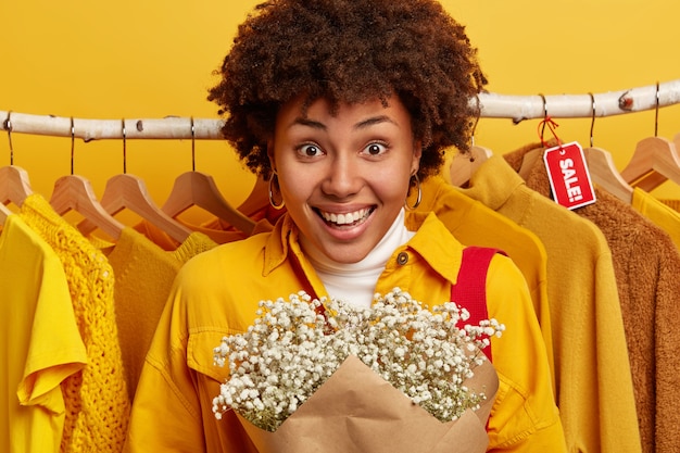 Photo gratuite consommateur femme bouclée aime faire du shopping, se tient avec bouquet dans une boutique de mode.