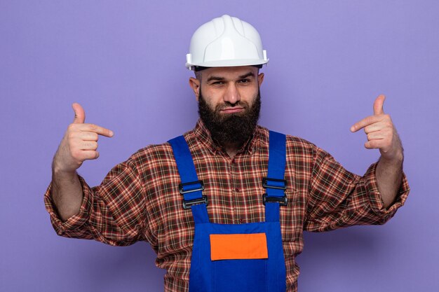 Constructeur barbu en uniforme de construction et casque de sécurité à la confiance souriante pointant sur lui-même debout sur fond violet