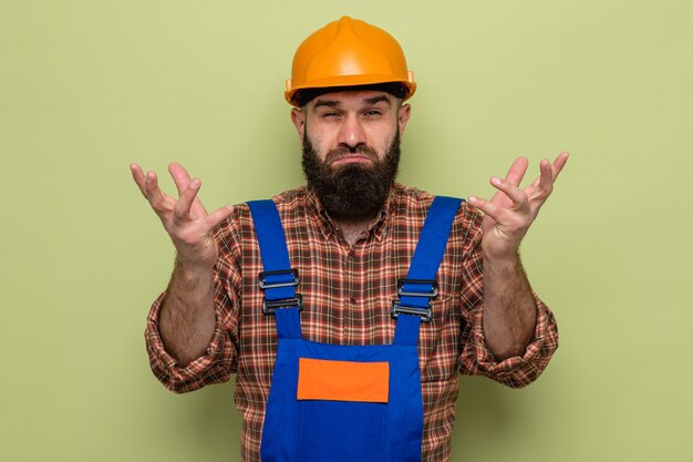 Constructeur barbu en uniforme de construction et casque de sécurité regardant la caméra confus haussant les épaules debout sur fond vert