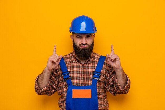 Constructeur barbu en uniforme de construction et casque de sécurité regardant avec un visage sérieux pointant l'index vers le haut