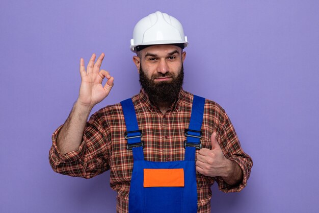 Constructeur barbu en uniforme de construction et casque de sécurité à sourire gaiement faisant signe ok montrant les pouces vers le haut