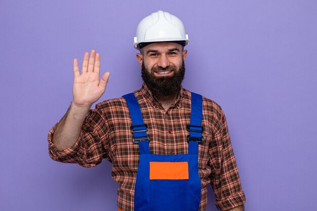 Constructeur barbu en uniforme de construction et casque de sécurité à sourire gaiement montrant cinquième avec palm