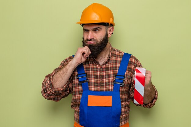 Constructeur barbu en uniforme de construction et casque de sécurité tenant du ruban adhésif regardant de côté avec une expression pensive sur la pensée du visage