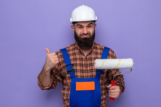 Constructeur barbu en uniforme de construction et casque de sécurité tenant un rouleau à peinture à sourire confiant montrant les pouces vers le haut