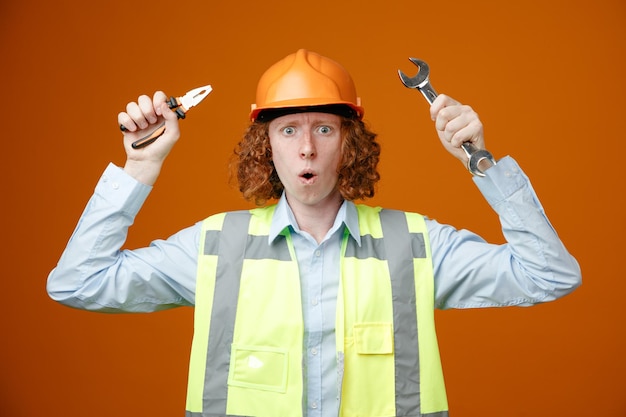 Constructeur jeune homme en uniforme de construction et casque de sécurité tenant une clé et une pince regardant la caméra étonné et surpris debout sur fond orange