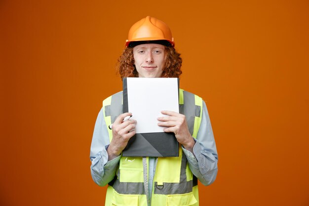 Constructeur jeune homme en uniforme de construction et casque de sécurité tenant presse-papiers et marqueur regardant la caméra souriant confiant debout sur fond orange