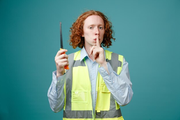 Constructeur jeune homme en uniforme de construction tenant une scie regardant la caméra avec un visage sérieux faisant un geste de silence avec le doigt sur les lèvres debout sur fond bleu