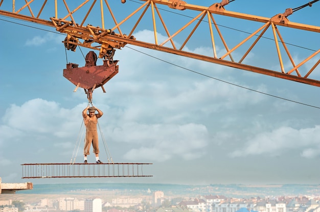 Constructeur musclé avec torse nu debout sur une construction en fer en hauteur et tenant par des cordes. Homme portant un chapeau et des vêtements de travail en regardant la caméra. Ciel bleu avec des nuages en arrière-plan.