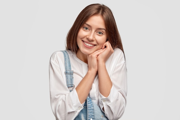 Content jeune femme garde les mains près de la joue, sourit positivement, exprime le bonheur et les sentiments véridiques, vêtue d'une tenue décontractée