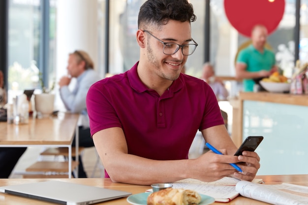 Contenu jeune blogueur masculin tient un téléphone portable, télécharge des fichiers, envoie des messages texte, utilise une connexion Internet gratuite, pose contre l'intérieur du café, recherche des informations. Concept de technologie et de travail