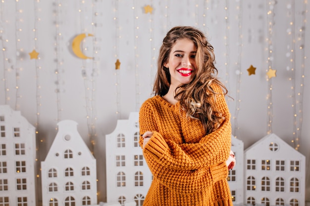 Contre des décorations chaudes en carton en forme de guirlandes et de maisons blanches, belle femme aux cheveux bruns en beau pull chaud posant