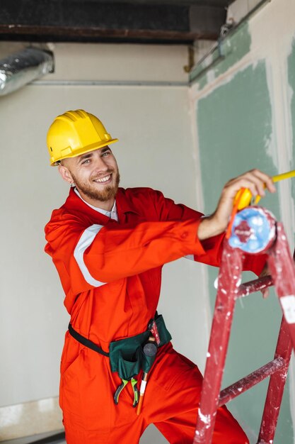 Contremaître souriant en vêtements de travail orange et casque jaune debout sur une échelle avec un ruban à mesurer à la main regardant joyeusement à huis clos sur le travail