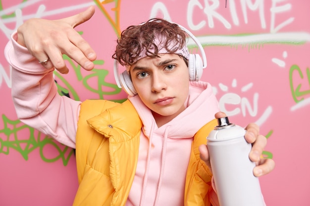 Cool beau adolescent vêtu d'un sweat à capuche et d'un gilet détient un spray aérosol fait des graffitis créatifs sur les murs de la rue écoute de la musique via des écouteurs sans fil