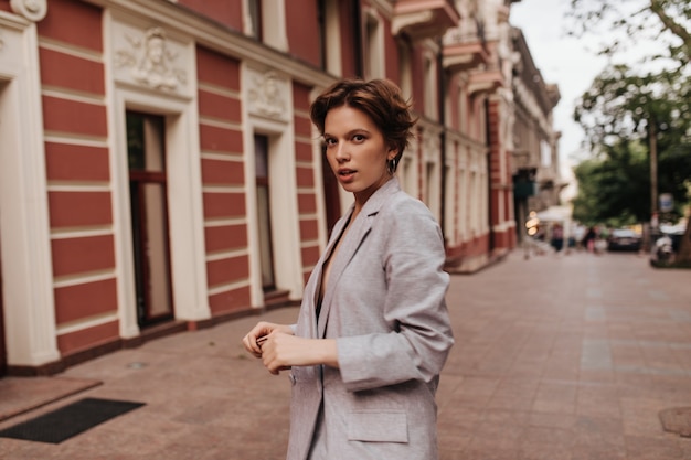 Cool femme en costume gris se penche sur la caméra à l'extérieur. Portrait de jeune fille aux cheveux courts en veste surdimensionnée se promenant dans la ville