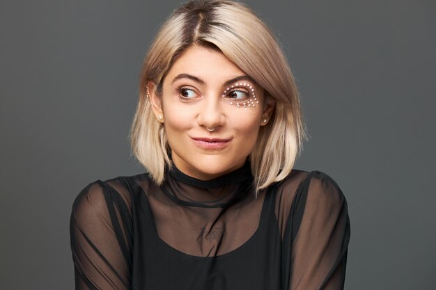 Cool jeune femme de race blanche à la mode avec une coupe de cheveux élégante et un piercing facial pensant à quelque chose d'agréable, faisant des plans, ayant une bonne idée, regardant ailleurs avec un sourire énigmatique mystérieux