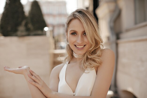 Cool joyeuse femme blonde en robe blanche à col en v et collier de perles sourit sincèrement, regarde dans l'appareil photo et pointe vers la gauche dans la rue