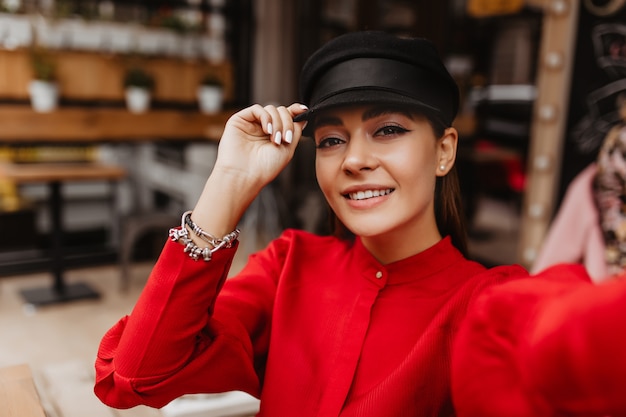 Cool selfie sur fond de café de rue. Jeune fille vêtue d'un élégant costume de soie rouge avec des bracelets en argent et des boucles d'oreilles en forme de perles sourit