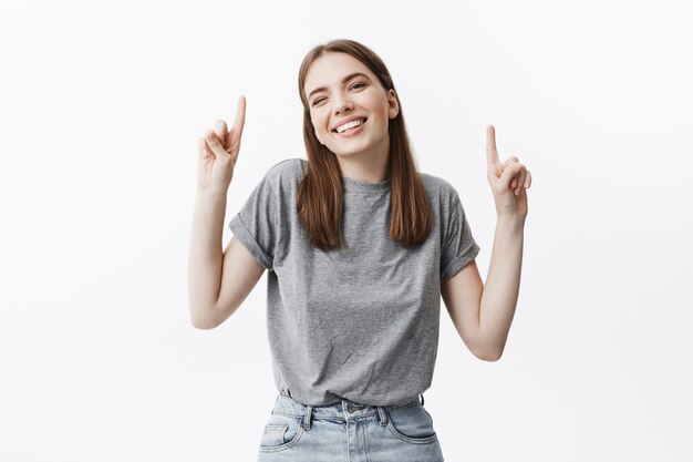 Copiez l'espace. Portrait de joyeuse belle étudiante brune souriante avec des dents, avec une expression satisfaite et excitée, pointant à l'envers avec les doigts sur les deux mains.