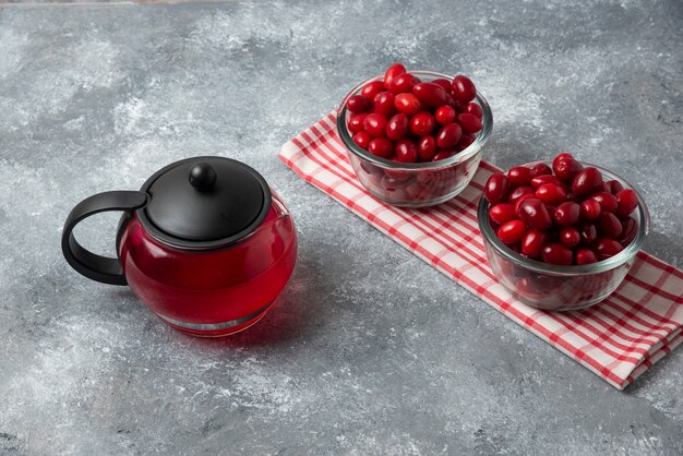 Photo gratuite cornels rouges dans la tasse en verre sur une serviette à carreaux avec une bouilloire de jus.