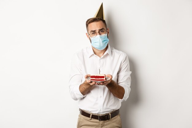 Coronavirus, quarantaine et jours fériés. Homme triste ne peut pas souffler la bougie du gâteau d'anniversaire, portant un masque facial et se plaignant, debout