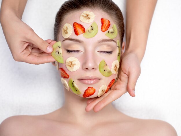 Cosméticienne faisant un masque de fruits sur un visage de belle jeune femme - à l'intérieur