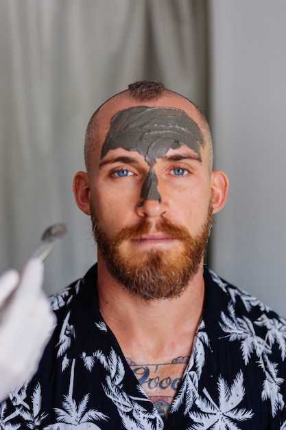 Photo gratuite cosmétologue appliquer un masque d'argile avec une brosse sur le visage de jeune homme beau dans une clinique de beauté