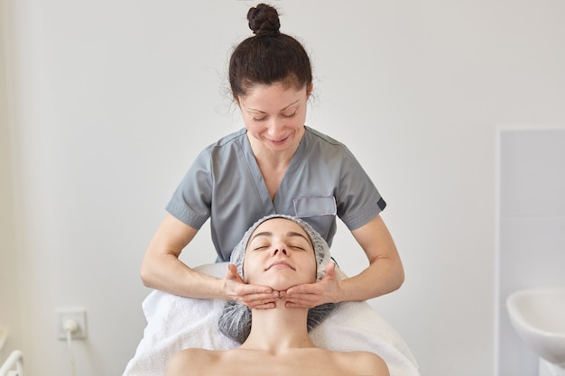 La cosmétologue porte une robe grise massant le visage du patient.