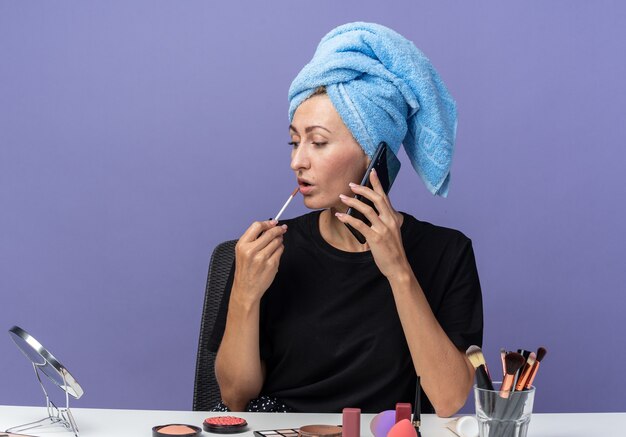 À côté de la belle jeune fille assise à table avec des outils de maquillage essuyant les cheveux dans une serviette parle au téléphone en appliquant un brillant à lèvres isolé sur un mur bleu