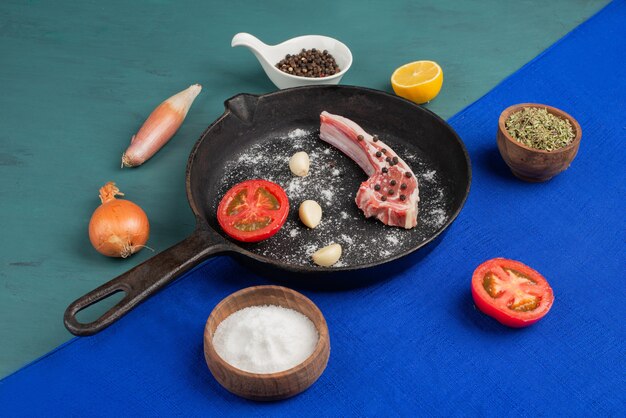 Côte crue dans une poêle noire avec des légumes et des épices sur table bleue.