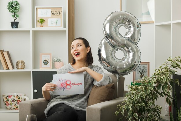 Côté joyeux, belle fille le jour de la femme heureuse tenant une carte de voeux assise sur un fauteuil dans le salon