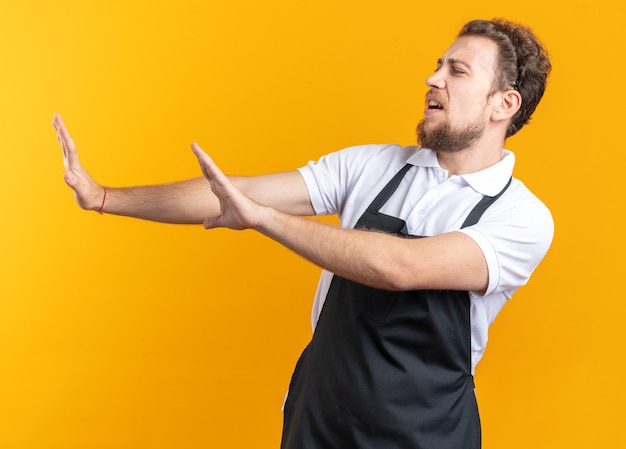 À Côté De Mécontent Jeune Homme Barbier En Uniforme Tenant Les Mains à Côté Isolé Sur Fond Jaune