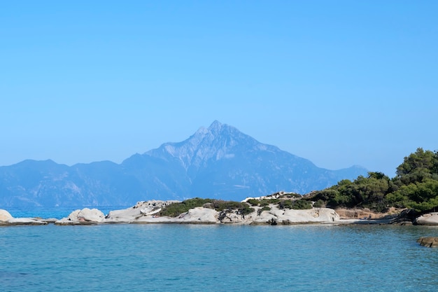 Côte de la mer Égée avec des rochers et des gens au repos, montagne au loin, verdure au premier plan, eau bleue, Grèce