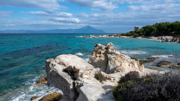 Côte de la mer Égée avec verdure autour, rochers, buissons et arbres, eau bleue avec vagues, montagne