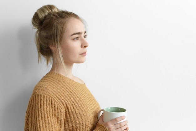 Sur Le Côté, Sérieuse Jeune Femme Réfléchie Avec Une Coiffure En Désordre Tenant Une Grande Tasse, Boire Du Cacao Chaud à La Maison, Debout Au Mur Blanc