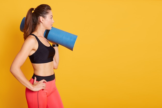 Côté wiev de jeune belle femme de race blanche debout et tenant un tapis de yoga bleu sur l'épaule, étant prêt pour l'exercice dans la salle de gym