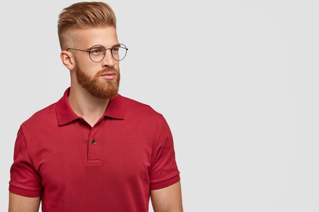 Coup de côté d'un homme au gingembre avec une coupe de cheveux à la mode, une barbe épaisse, concentré de côté, pense à un nouveau projet, porte un t-shirt rouge décontracté brillant, isolé sur un mur blanc