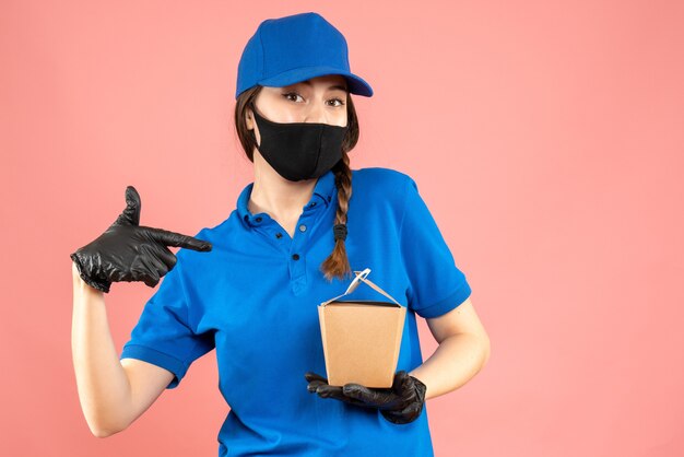 Coup de demi-corps d'une coursière confiante portant un masque médical et des gants tenant une petite boîte sur fond de pêche pastel