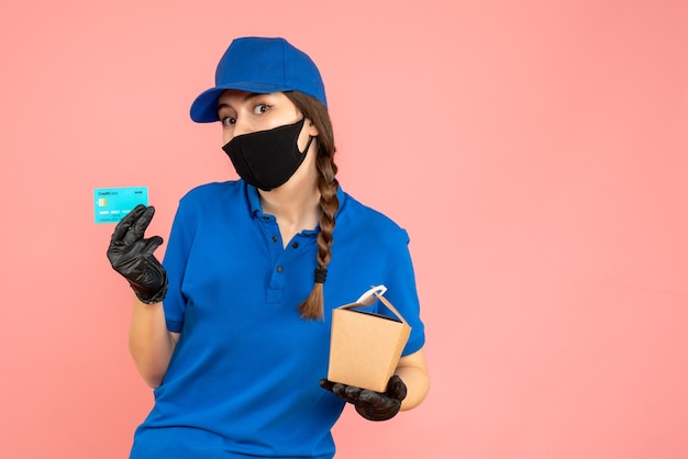 Coup de demi-corps d'une coursière portant un masque médical et des gants tenant une petite boîte et une carte bancaire sur fond de pêche pastel