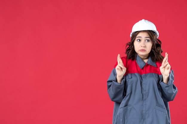 Coup de demi-corps d'une femme builder surprise en uniforme avec un casque et croisant les doigts sur fond rouge isolé