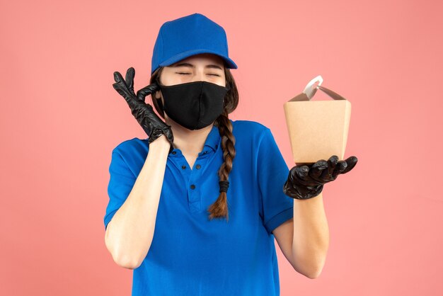 Coup de demi-corps d'une fille de courrier nerveux portant un masque médical et des gants tenant une petite boîte sur fond de pêche pastel