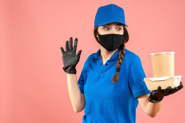 Coup de demi-corps d'une fille de courrier se demandant portant un masque médical et des gants tenant une petite boîte de café sur fond de pêche pastel