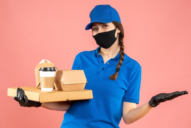 Coup de demi-corps d'une fille de messagerie satisfaite portant un masque médical et des gants tenant des commandes sur fond de pêche pastel