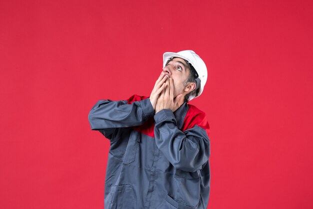 Coup de demi-corps d'un jeune constructeur choqué en uniforme avec un casque en levant sur un mur rouge isolé