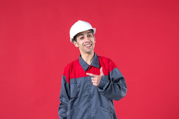 Coup de demi-corps d'un jeune constructeur nerveux en uniforme avec un casque de protection montrant quelque chose sur le côté droit sur un mur rouge isolé