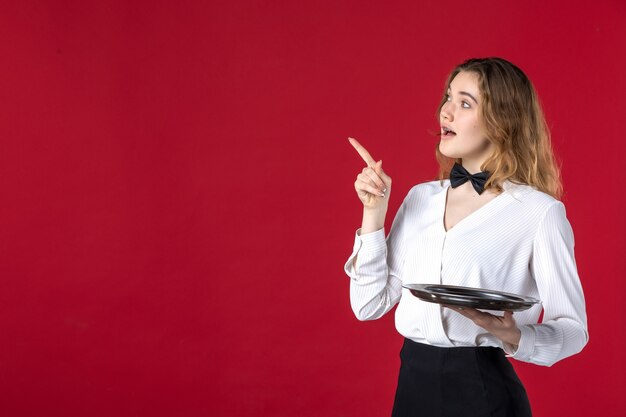 Coup de demi-corps d'une serveuse curieuse papillon sur le cou et tenant un plateau pointant vers le haut sur fond rouge